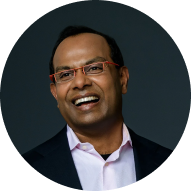 Professional headshot: smiling man in a dark blazer and pink shirt against a dark teal background.