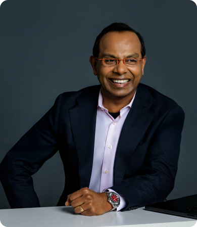 Smiling man in a navy blazer and pink shirt sits at a desk with a laptop nearby, hands folded on the table.