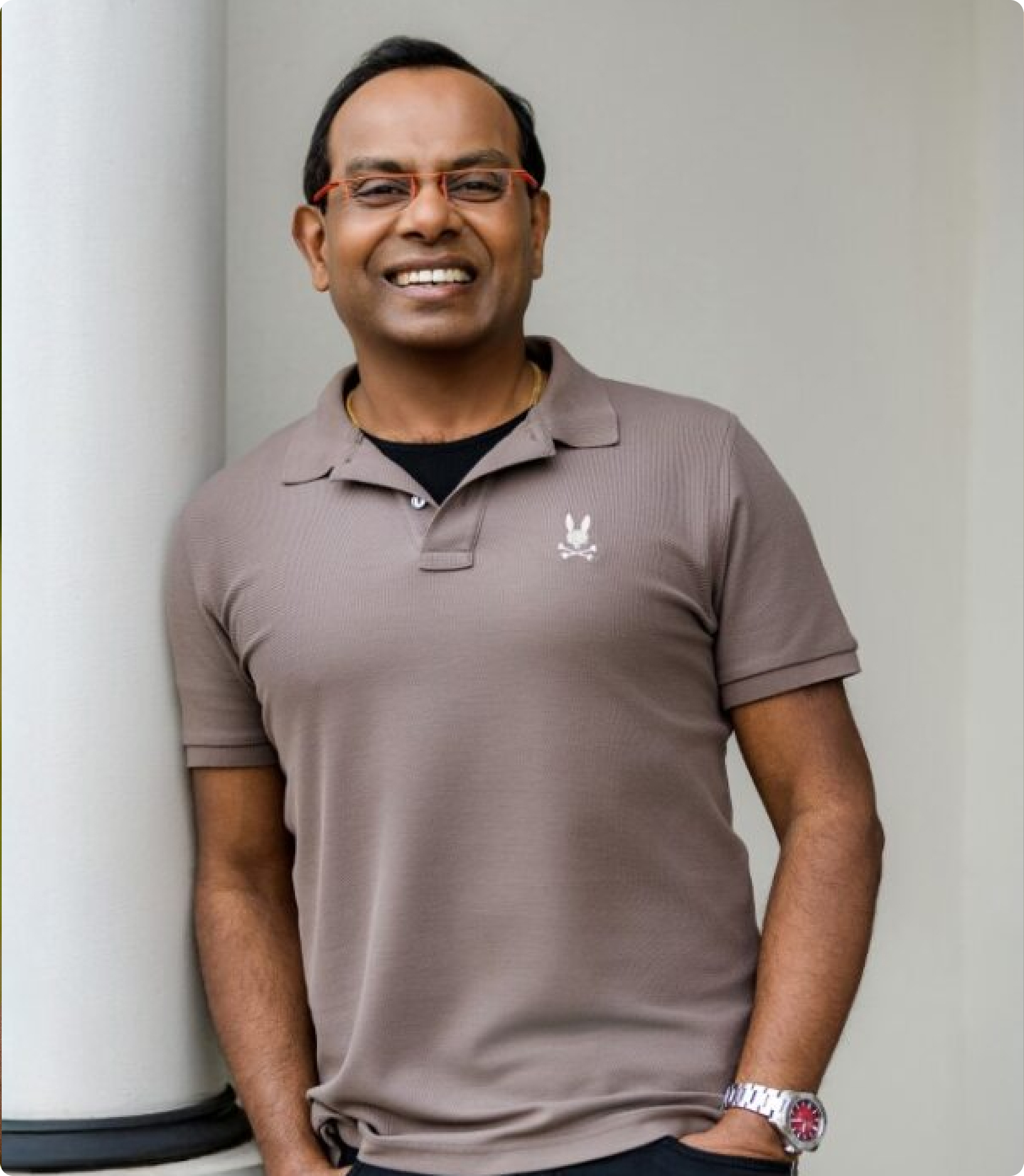 Smiling man wearing a brown polo shirt with a logo and red glasses, standing beside a light pillar against a neutral background.