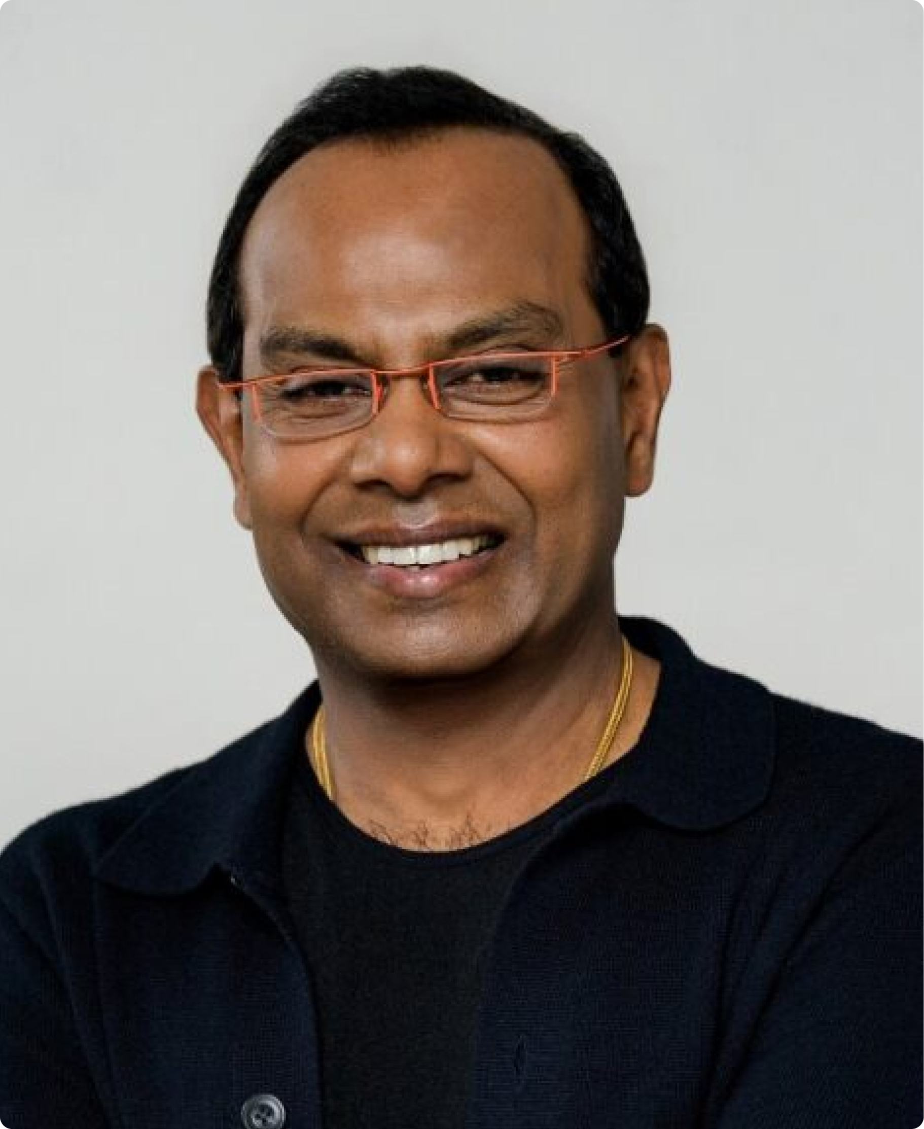 Smiling man wearing orange-framed glasses and a black polo against a light gray background.