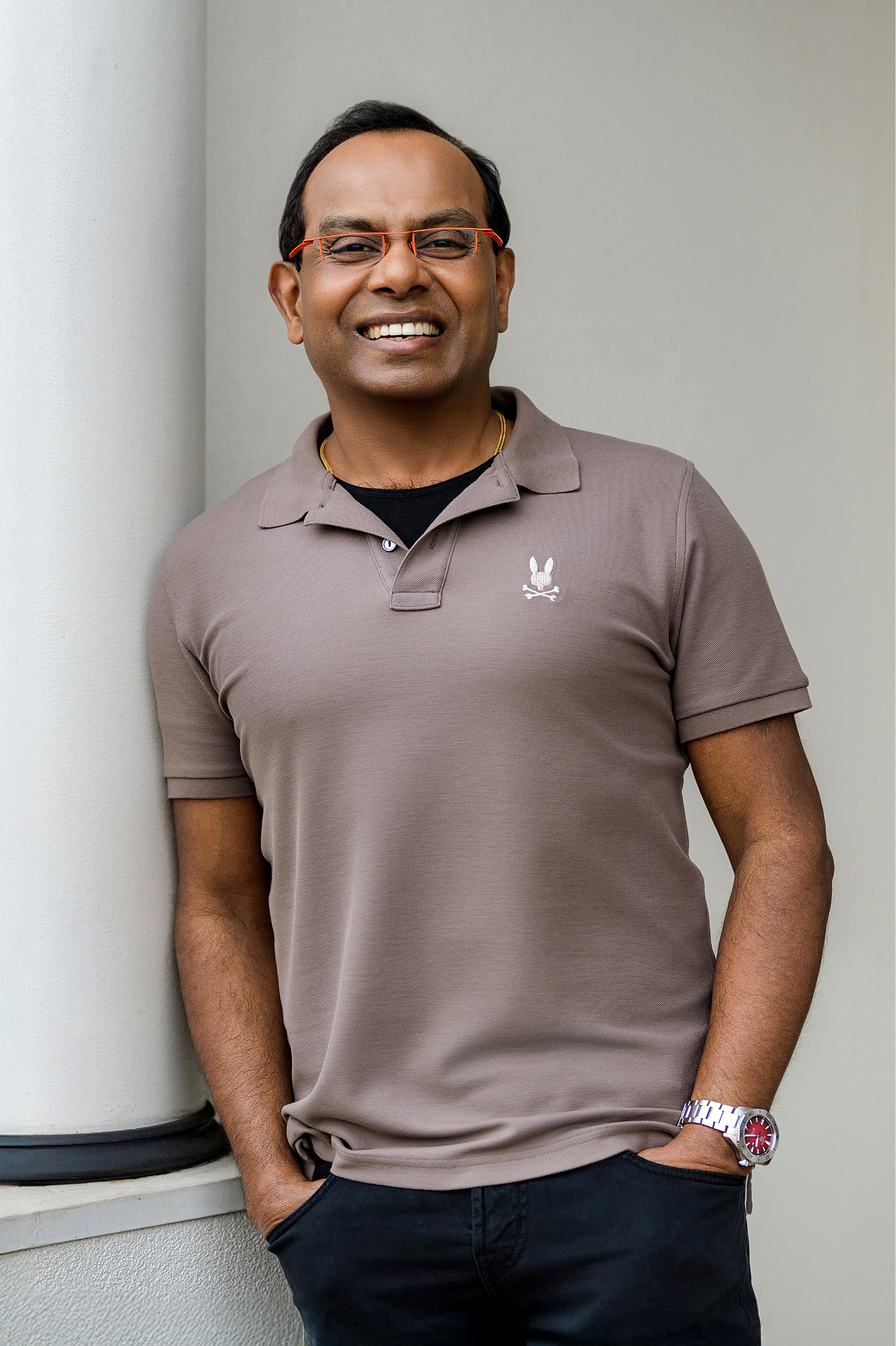 Smiling man in a taupe polo shirt leaning against a light column, hands in pockets.