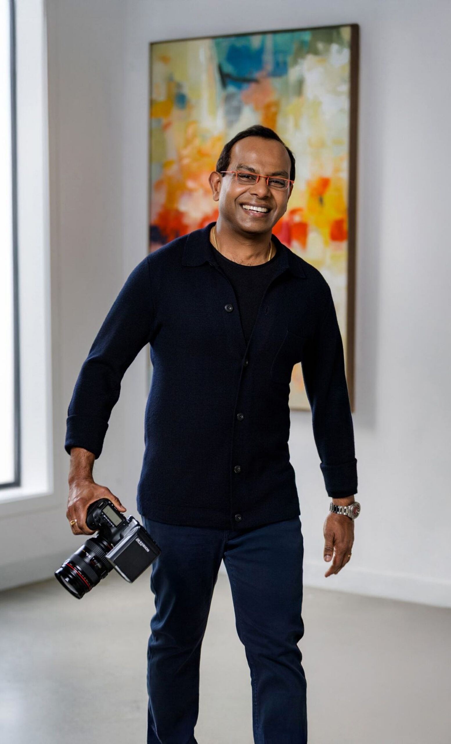 Photographer smiling in a navy cardigan, holding a professional camera indoors with a colorful abstract painting in the background.