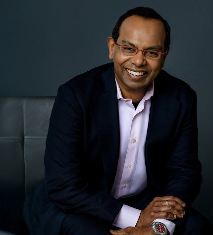 Smiling man in a dark blazer and light shirt sits on a gray couch, hands folded, posing for a professional portrait.