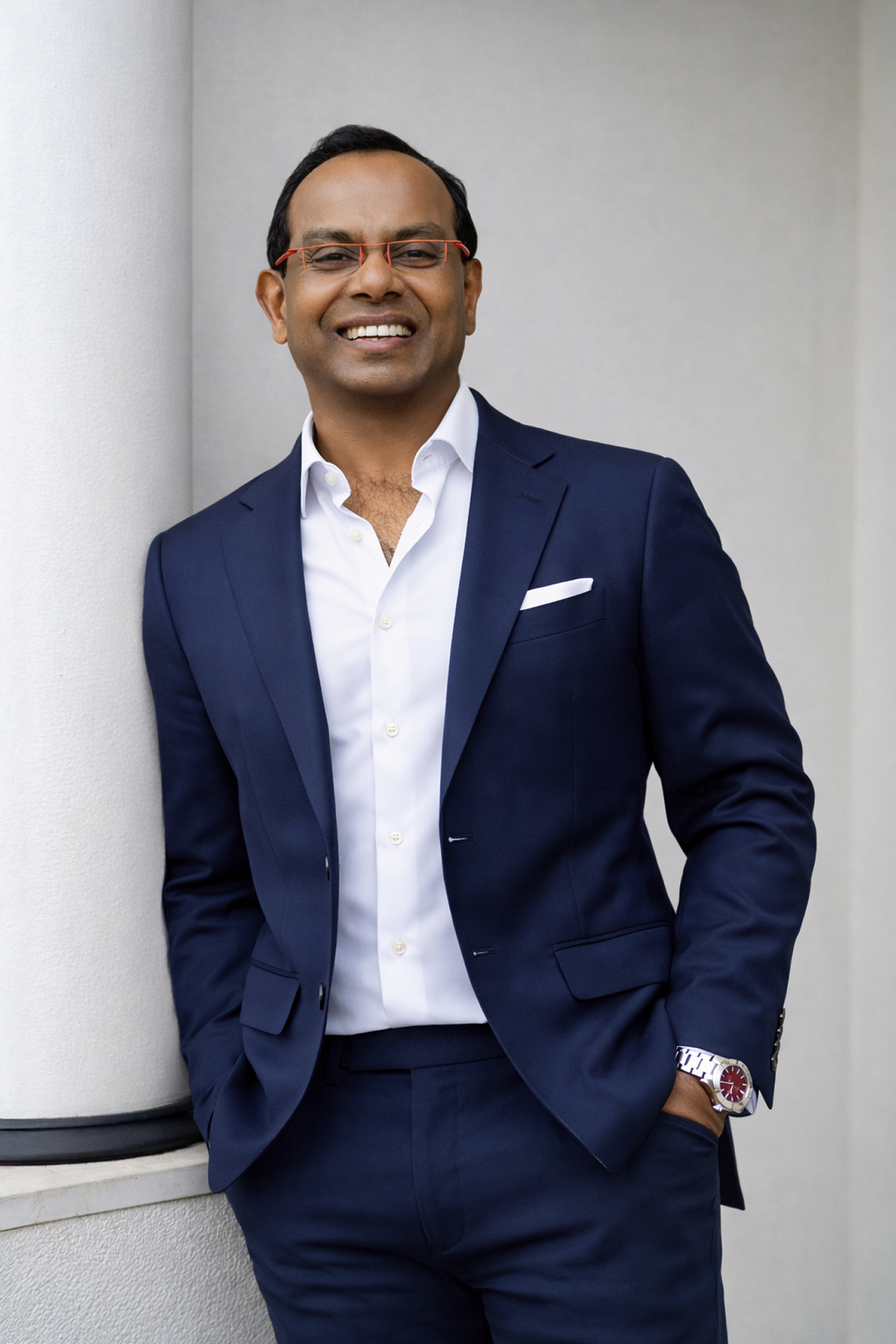 Smiling man in a navy suit stands against a light column, wearing glasses and a white shirt.