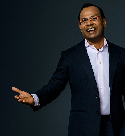 Smiling man in a dark suit and pink shirt extending his right arm as if presenting.