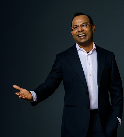 Smiling man in a dark suit and pink shirt extends his right hand in a welcoming or presenting gesture.