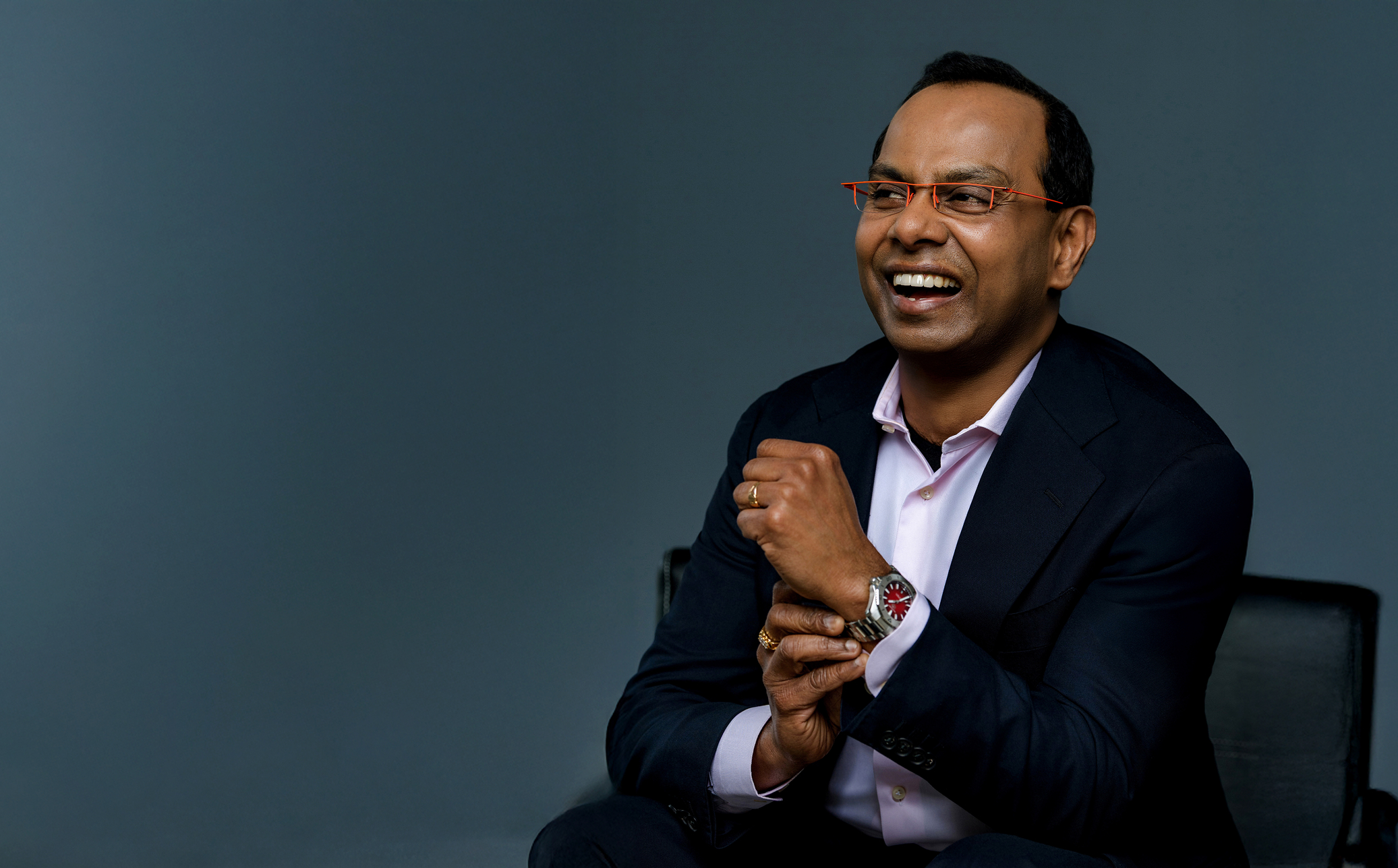 Smiling man in a navy suit and pink shirt sits against a blue backdrop, laughing groovily.