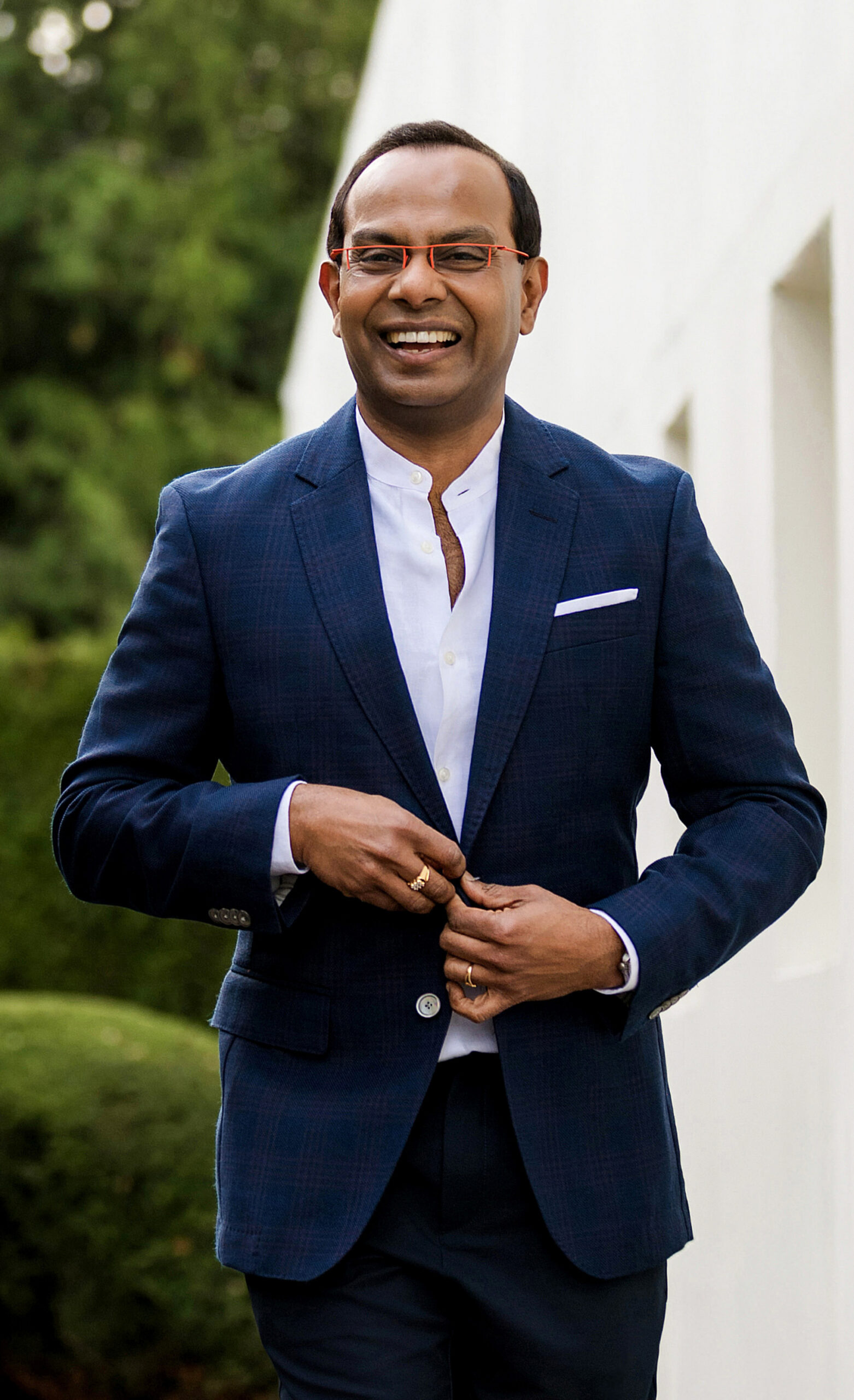 Smiling man in a blue suit and white shirt outdoors beside a white wall.